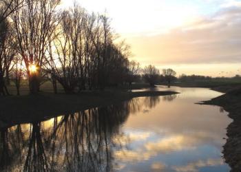Ein kleiner Fluss umgeben von Bäumen, die Sonne geht gerade auf und spiegelt sich im Wasser