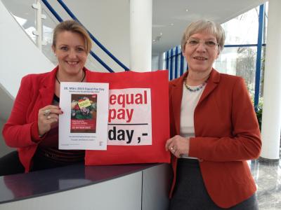 Zwei Frauen schauen lächelnd in die Kamera und halten Plakate und Taschen für den Equal Pay Day 2015 hoch 