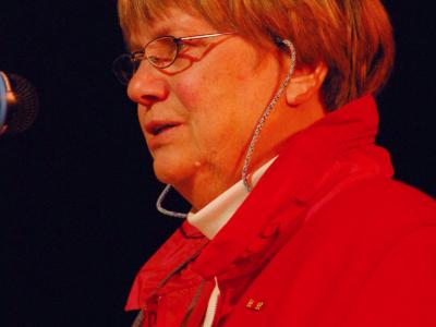 Eine Frau mit roter Jacke und Brille steht vor einem Mikrofon 